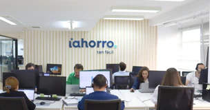 Varios trabajadores en sus puestos de trabajo, al fondo, el logo de iAhorro en una pared.